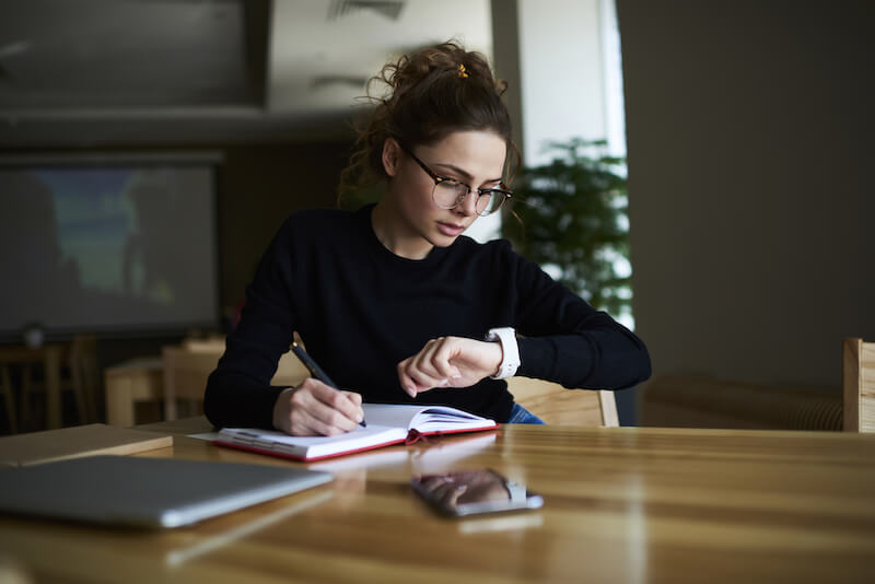 Timeblocking. a woman staring at the time on her watch while writing in a book.